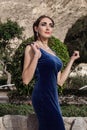 Beautiful young brunette woman in a velvet blue evening dress with a clutch, posing in nature.