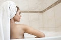 Beautiful young brunette woman wearing white towel on her head sitting in bath half a turn in bright bathroom