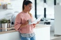 Beautiful young business woman drinking a cup of coffee while using her smart phone while holding papers in the kitchen at home Royalty Free Stock Photo