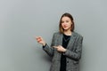 Beautiful young business woman in a jacket stands on a background of a gray wall and shows a finger to the side on copy space with Royalty Free Stock Photo