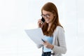 Beautiful young business woman using her mobile phone and reviewing papers in the office. Royalty Free Stock Photo