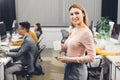 Beautiful young businesswoman with cup and smartphone smiling at camera while colleagues working behind Royalty Free Stock Photo