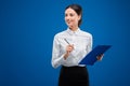 Beautiful, young businesswoman looking happily towards the left while writing on her clipboard,  on blue background Royalty Free Stock Photo