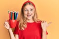 Beautiful young caucasian girl holding art colored pencils pointing thumb up to the side smiling happy with open mouth Royalty Free Stock Photo