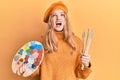 Beautiful young caucasian girl holding paintbrush and painter palette angry and mad screaming frustrated and furious, shouting Royalty Free Stock Photo