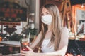 A beautiful young caucasian girl wearing a mask sits in a street cafe with drink. COVID-19 quarantine in the city