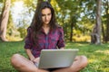 Beautiful young Caucasian happy woman wearing plaid shirt, sitting on grass in front of laptop pc, working on her laptop computer Royalty Free Stock Photo