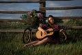 A beautiful young Caucasian smiling happy girl in purple dress in a wreath and with a guitar and blooming lilac in the