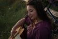 A beautiful young Caucasian smiling happy girl in purple dress in a wreath and with a guitar and blooming lilac in the