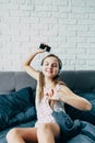Beautiful young Caucasian teenage girl sitting in bed with modern headphones is holding phone in her hand, dancing to cool music a Royalty Free Stock Photo