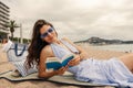 Beautiful young caucasian woman reading guidebook resting on beach with city in background.