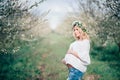 Beautiful young cheerful pregnant woman in wreath of flowers on head touching belly while walking in spring tree garden. Beauty