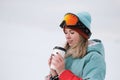 A beautiful young chewing woman in an extreme sports suit, hat and helmet with a mask on the winter slope is holding a Royalty Free Stock Photo