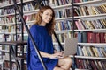 Beautiful young college student sitting on stairs in the library, working on laptop. Woman wearing blue dress, huge bookshelf