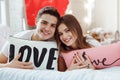 Beautiful young couple in bedroom is sitting on bed, smiling and looking at camera. Celebrating Saint Valentine`s Day Royalty Free Stock Photo