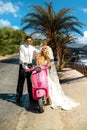 Beautiful young couple, blonde female model with her husband in amazing wedding clothes sitting on scooter