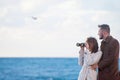 Happy beautiful young couple fashionable man and woman in autumn coat looking through binoculars near sea with horizon and flying Royalty Free Stock Photo