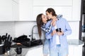 Beautiful young couple is hugging and smiling while drinking tea or coffee in kitchen at home. Man is kissing his charming girl Royalty Free Stock Photo