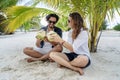 A beautiful young couple joyfully sits on white sand under a palm tree with coconuts in their hands on the seashore under a green Royalty Free Stock Photo