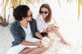 A beautiful young couple joyfully sits on white sand under a palm tree with coconuts in their hands on the seashore under a green Royalty Free Stock Photo