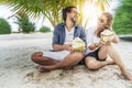 A beautiful young couple joyfully sits on white sand under a palm tree with coconuts in their hands on the seashore under a green Royalty Free Stock Photo