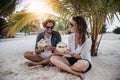 A beautiful young couple joyfully sits on white sand under a palm tree with coconuts in their hands on the seashore under a green Royalty Free Stock Photo