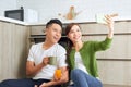 Beautiful young couple in love sitting on the kitchen floor, taking selfies using smart phone Royalty Free Stock Photo