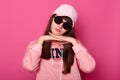 Beautiful young dark haired teenager with big black sunglasses, wearing pink hoody and cap, looks upset, poses over empty pink Royalty Free Stock Photo