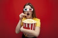 Beautiful young emotional girl in 3D glasses eats popcorn, watches a movie and screams on a red color background, surprised viewer Royalty Free Stock Photo