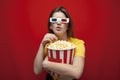 Beautiful young emotional girl in 3D glasses eats popcorn, watches a movie and screams on a red color background, surprised viewer Royalty Free Stock Photo