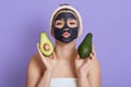 Beautiful young European woman with black facial mask and fresh avocado in hands isolated on lilac background, making kissing Royalty Free Stock Photo