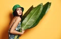 Beautiful young fashion girl with perfect skin in green hat hold tropical banana leaf in hands and covers Royalty Free Stock Photo