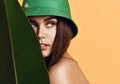 Beautiful young fashion girl with perfect skin in green hat hold tropical banana leaf in hands and covers Royalty Free Stock Photo