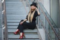 Beautiful young fashionable woman posing in black suite, red shoes with high heels and black hat. Vogue style. Urban background