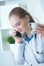 Beautiful young female doctor sitting in front of working table with laptop and documents on it calling on the phone and Royalty Free Stock Photo