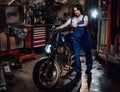 Beautiful young female mechanic in blue overalls posing with custom bobber in garage or workshop Royalty Free Stock Photo