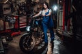 Beautiful young female mechanic in blue overalls posing with custom bobber in garage or workshop Royalty Free Stock Photo