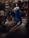 Beautiful young female mechanic in blue overalls posing with custom bobber in garage or workshop Royalty Free Stock Photo