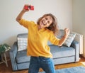 Beautiful young female in a yellow shirt is dancing happy and joyful in the living room with a smartphone in her hands, emotion Royalty Free Stock Photo