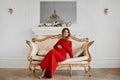 Beautiful young gestation woman in fashionable red evening dress sits on the gold vintage couch at luxury interior