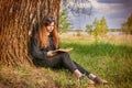 Beautiful young girl in dark clothes, sitting on the green grass near a large tree in the Park by the lake and reading a book unde Royalty Free Stock Photo