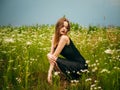 Beautiful young girl in a black evening dress poses crouching in front of a daisy field