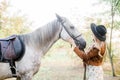 Beautiful young girl with blond hair in a suede jacket with a fringe , wearing black floppy hat, smiling and stroking her horse Royalty Free Stock Photo