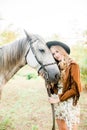 Beautiful young girl with blond hair in a suede jacket with a fringe , wearing black floppy hat, smiling and stroking her horse Royalty Free Stock Photo