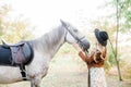 Beautiful young girl with blond hair in a suede jacket with a fringe , wearing black floppy hat, smiling and stroking her horse Royalty Free Stock Photo