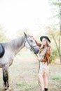 Beautiful young girl with blond hair in a suede jacket with a fringe , wearing black floppy hat, smiling and stroking her horse Royalty Free Stock Photo