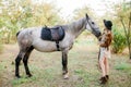 Beautiful young girl with blond hair in a suede jacket with a fringe , wearing black floppy hat, smiling and stroking her horse Royalty Free Stock Photo