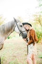 Beautiful young girl with blond hair in a suede jacket with a fringe , wearing black floppy hat, smiling and stroking her horse Royalty Free Stock Photo