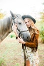 Beautiful young girl with blond hair in a suede jacket with a fringe , wearing black floppy hat, smiling and stroking her horse Royalty Free Stock Photo