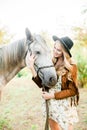 Beautiful young girl with blond hair in a suede jacket with a fringe , wearing black floppy hat, smiling and stroking her horse Royalty Free Stock Photo